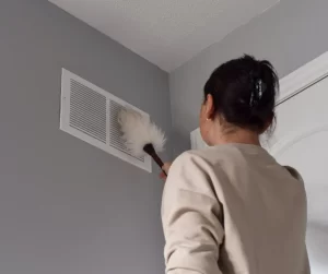 October Home Checklist. A woman cleaning her vents with a white feather duster.