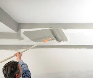 The Difference Between Ceiling Paint and Wall Paint. A guy painting the ceiling with a long paint roller.