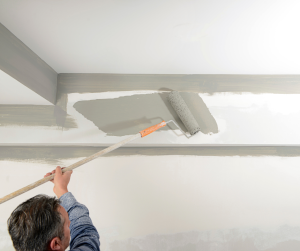 The Difference Between Ceiling Paint and Wall Paint. A guy painting the ceiling with a long paint roller.
